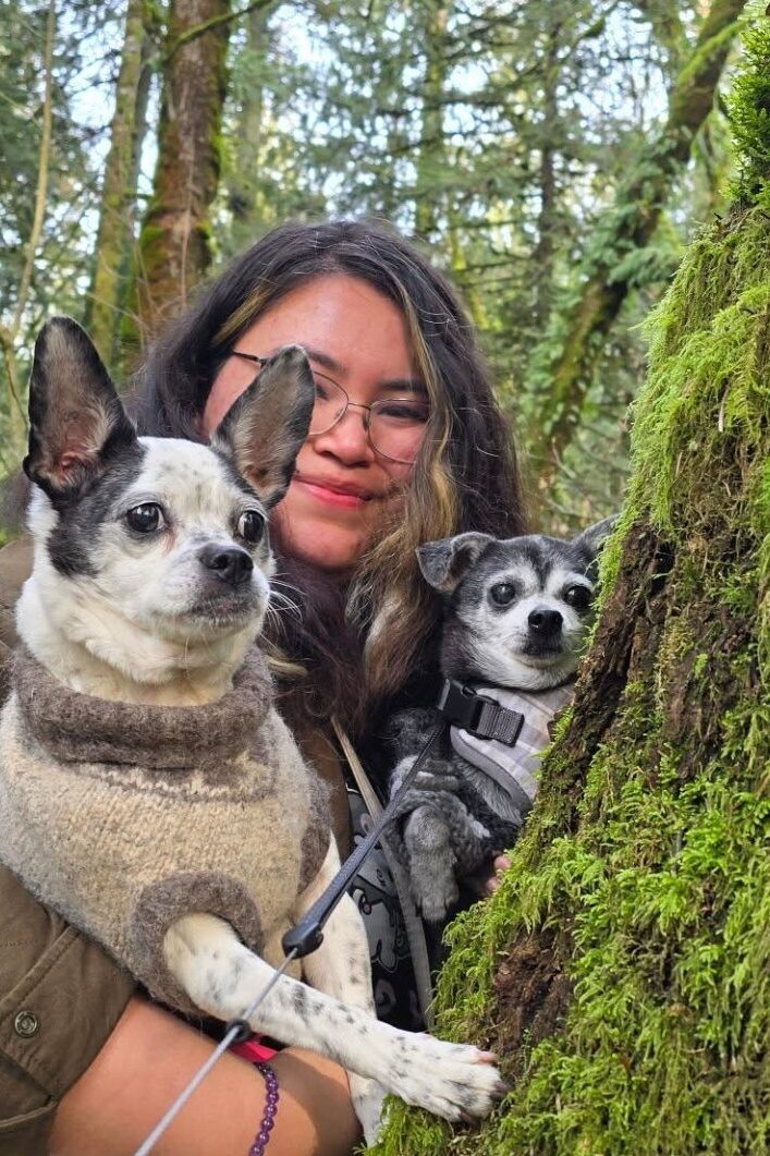 Bunny and Maeson are hoisted up against a tree, by Sierra-a coast salish women with tan skin and medium wavy length hair, who is smiling at the camera. Both dog's are wearing sweaters as they pose for the picture. Bunny is looking at something else in the distance, while Maeson is looking slightly to his left of the camera lens